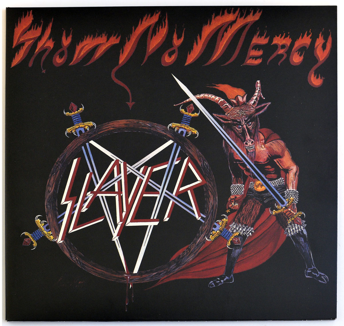 Album cover of Show No Mercy by Slayer. The artwork features a dark fantasy-inspired scene with a muscular, goat-headed demon warrior wielding a sword. He wears spiked boots, studded wristbands, and a belt that says “HEAVY.” The background is black with fiery red lettering spelling out the album title in a jagged, flame-like font. To the left, Slayer’s logo is displayed as a pentagram formed by interlocked swords, dripping with blood. The overall design is intense, evoking themes of darkness, power, and early thrash metal aesthetics.
